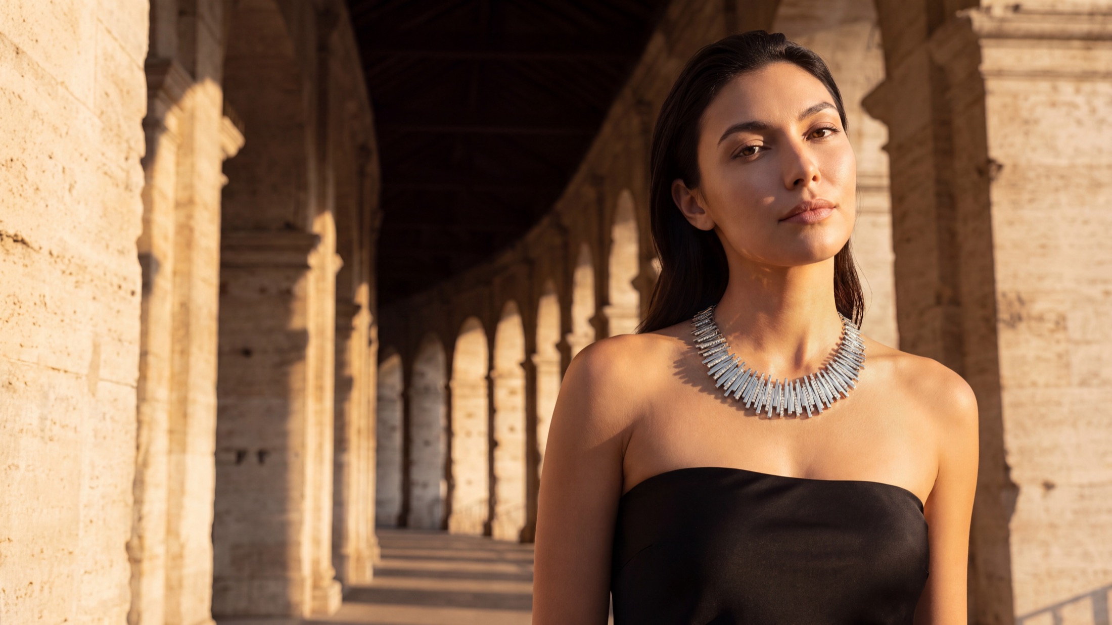 Model in a black dress wearing a diamond collar necklace under a stone arcade at golden hour